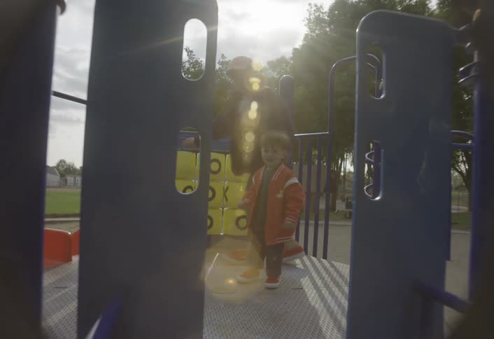 An older boy wearing a blue jacket plays with a toddler in a red jacket on the metal platform of a blue and yellow playground structure on a partly cloudy day interacting with a small orange ball and moving around as sunlight flares through the scene