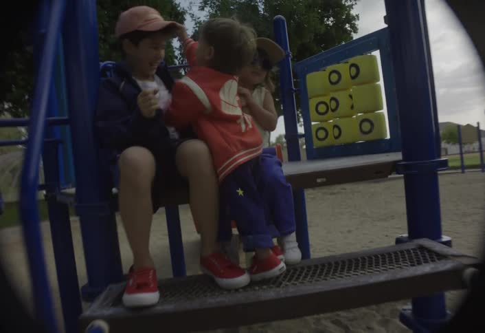 A boy wearing a baseball hat sits on a slide at the playground playing happily with two toddlers and having a fun time