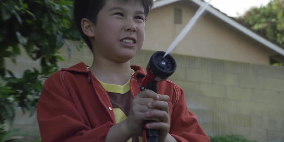 A young boy playfully aims a garden hose nozzle spraying a focused stream of water with a look of happy concentration on his face in a green backyard