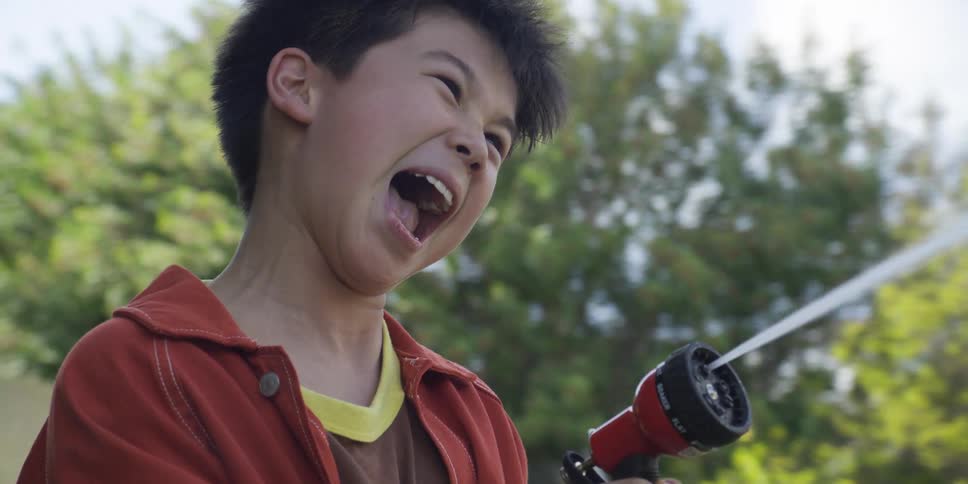 A young boy is shown outdoors likely in a yard holding a red and black hose nozzle that is spraying a stream of waterhe is wearing an orange or burntred jacket and has a wideopen mouth suggesting he is laughing screaming or shouting with excite