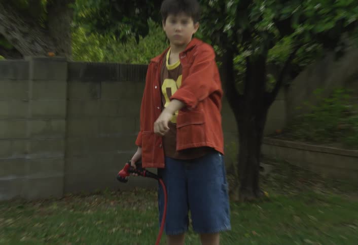 A young boy standing in a grassy yard dressed in an orange jacket and denim shorts holds a red garden hose nozzle and makes gestures toward the camera