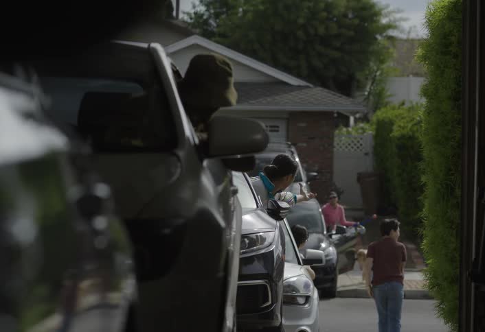 People lean out of parked cars arguing with each other in a suburban driveway lined with vehicles