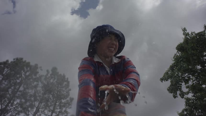 A smiling child wearing a hat and striped shirt gets splashed with water under a cloudy sky