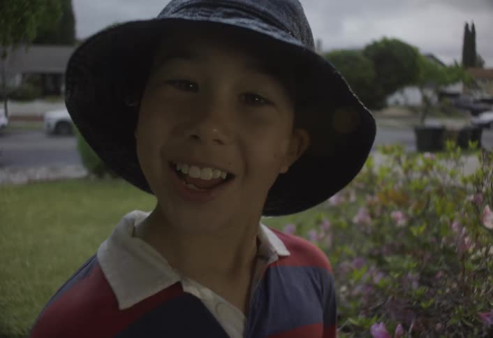 A boy with his face hidden by a bucket hat looks up suddenly breaking into a wide and joyful smile as the camera moves in close