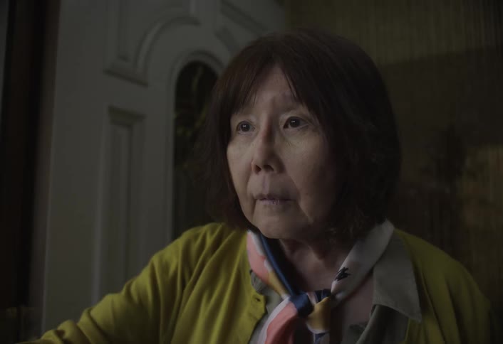 The video shows an older woman with shoulderlength dark hair and bangs looking slightly concerned or thoughtfulshe is wearing a yellow cardigan over a lightcolored shirt and a colorful patterned scarf is tied around her neck she appears to be st