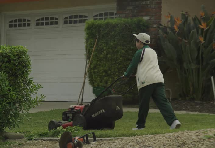 A boy pushes a lawn mower across the yard of a suburban home
