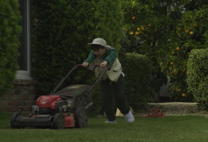 A child wearing a hat pushes a lawn mower across the backyard lawn at midday
