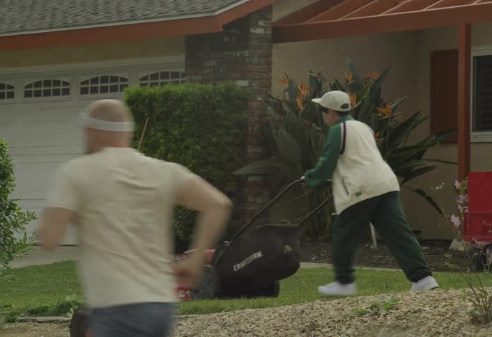 A young person in a cap and white vest pushes a craftsman lawnmower across the front lawn of a suburban home performing yard work