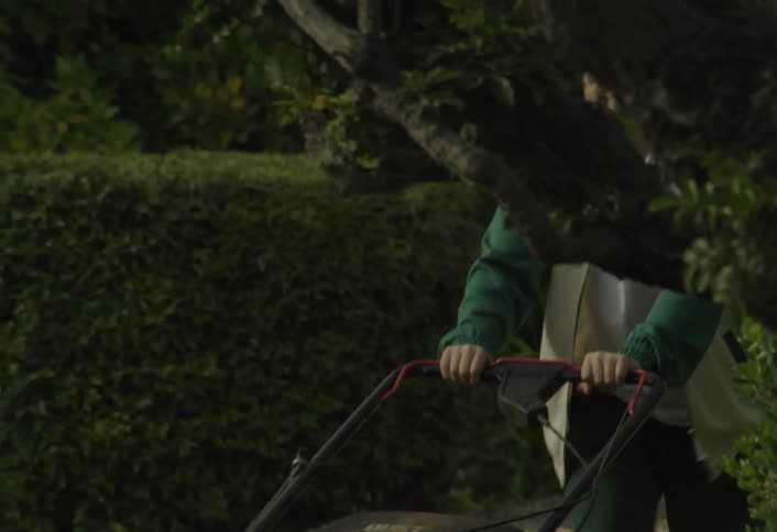 A young boy in a jacket and hat pushes a lawn mower across his yard navigating around trees and bushes as he diligently cuts the grass
