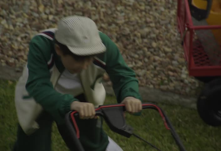 A boy is shown mowing a lawn with a walkbehind lawnmower the video cuts between closeup shots of the boy pushing the mower and wide overhead shots showing the yard a red wagon with tools and a car parked on the street