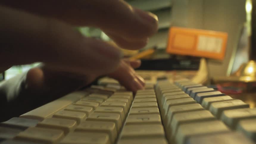 A closeup shot focuses on a persons hands typing on a mechanical keyboard with lightcolored keycaps the background is a dimly lit desk featuring a rotary phone blinds and a box