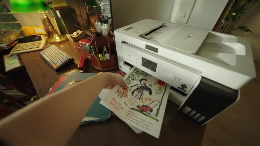 The first video shows a person taking a printed page featuring a brightly colored drawing of a man with a lawnmower off a desktop printer on a cluttered desk in an office space the second video shows a young boy in a yellow shirt posing making funny f