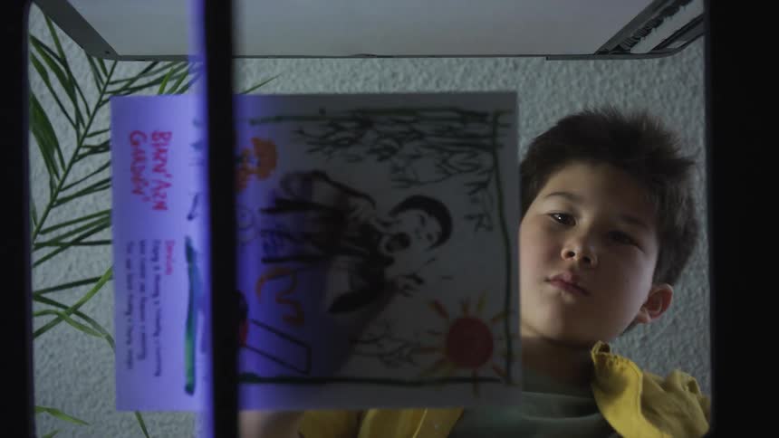 A young asian boy in a yellow garment puts an artwork against a lit surface to scan in an indoor setting