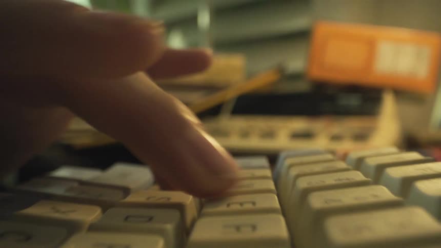 The video offers an extremely close lowangle view of hands rapidly typing on a lightcolored mechanical keyboard the scene is somewhat dark and blurred with the background vaguely showing desk items like a rotary phone and an orange box