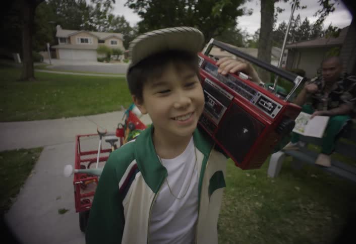 A young boy wearing a cap is holding a radio on his shoulder as he walks happily