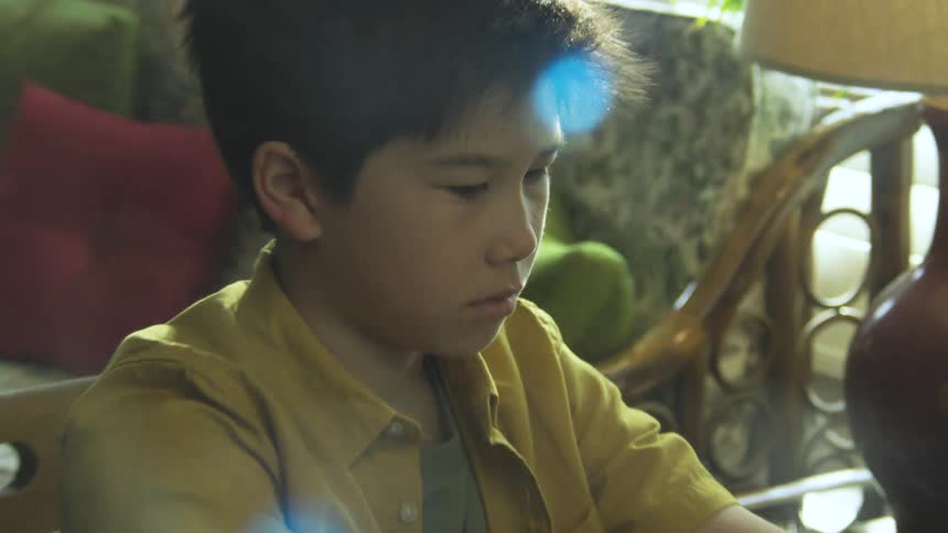 This video shows a young boy sitting in a room illuminated by sunlight coming through a windowthe camera alternates between medium shots of the boy and extreme closeups of his face and eyes conveying an intense focus or concentration as he looks dow