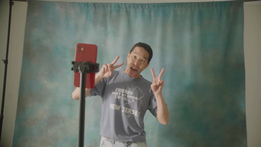 A man adjusts his smartphone on a stand and then records himself making a series of playful silly faces and peace signs in front of a blue backdrop
