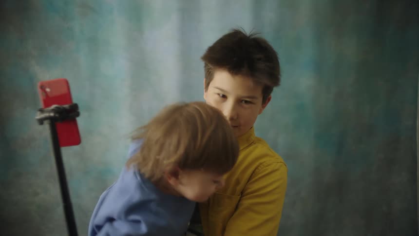 An older boy in a yellow shirt holds a smiling baby boy in a blue sweatshirt while the children look into a red smartphone mounted on a tripod against a blueandgray textured background