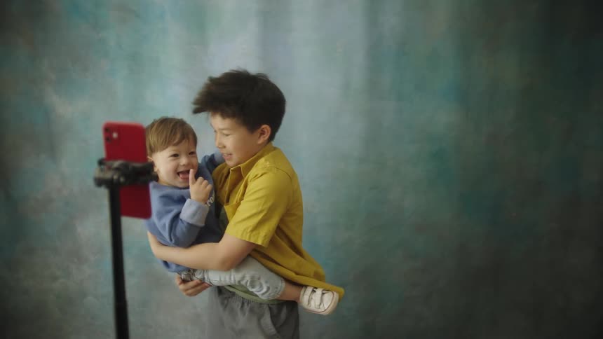 Two boys one older holding a younger child are standing in front of a bluegreen textured backdrop posing and making funny faces into a red smartphone mounted on a standthey are smiling and appear to be enjoying themselves while recording a video or
