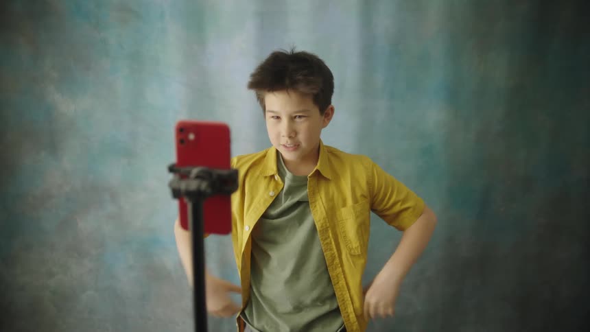 A cheerful boy in a yellow shirt jumps and strikes playful poses while recording a dance video on a red smartphone mounted against a textured blue studio backdrop