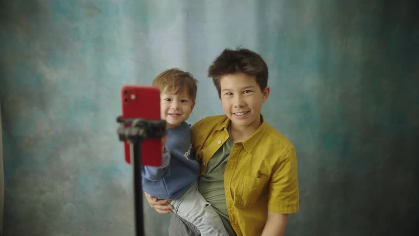 A boy is holding a toddler inside with a multi colored backround in front of a smart phone on a stand to take a selfie