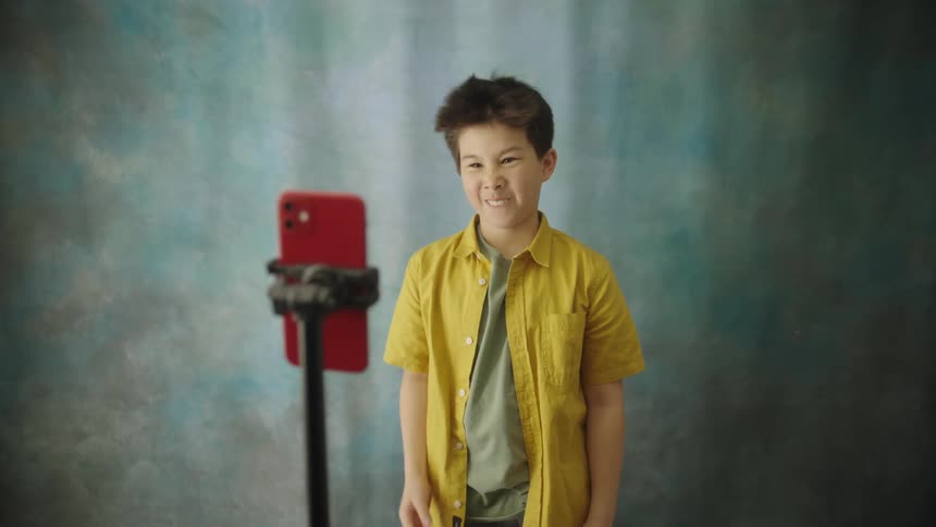 The video shows a young boy standing in front of a bluegreen textured backdrop recording himself with a red smartphone mounted on a tripod he poses and gestures to the camera starting with a thoughtful expression and hand on his chin then smiling wid