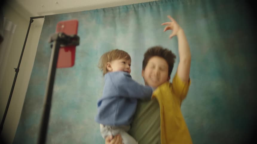An older boy in a yellow shirt holds a smiling toddler in a blue sweatshirt with a girl in a pink sweater too as they pose have fun and look into a red smartphone mounted on a tripod against a textured blue and gray backdrop