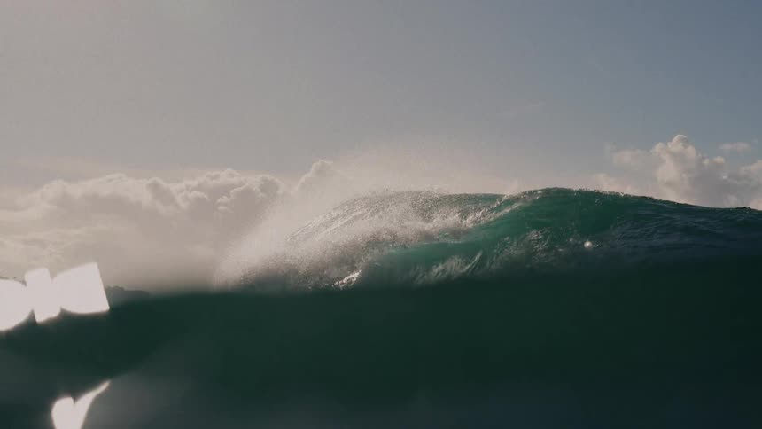 A dramatic lowangle shot captures a large ocean wave cresting and beginning to break with sunlight glinting through the spray the camera momentarily dips below the waters surface creating a hazy bright bokeh effect before the clip ends