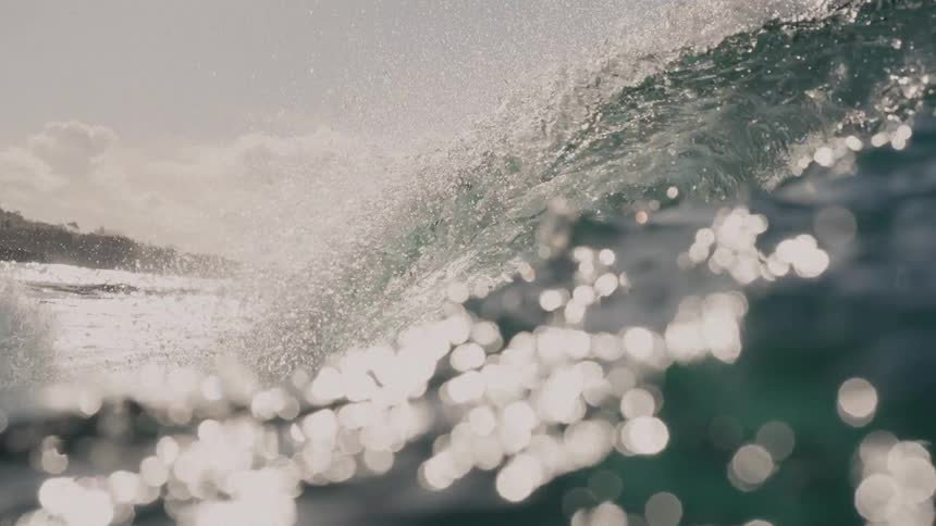 A massive ocean wave breaking and forming a barrel with sunlight intensely reflecting off the spraying water before the camera submerges into the deep green water