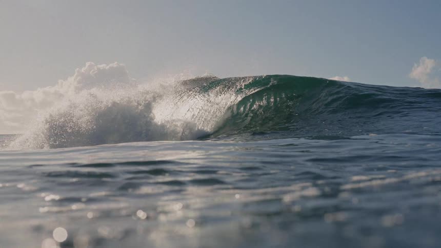 A powerful massive ocean wave breaks under a sunny partly cloudy sky