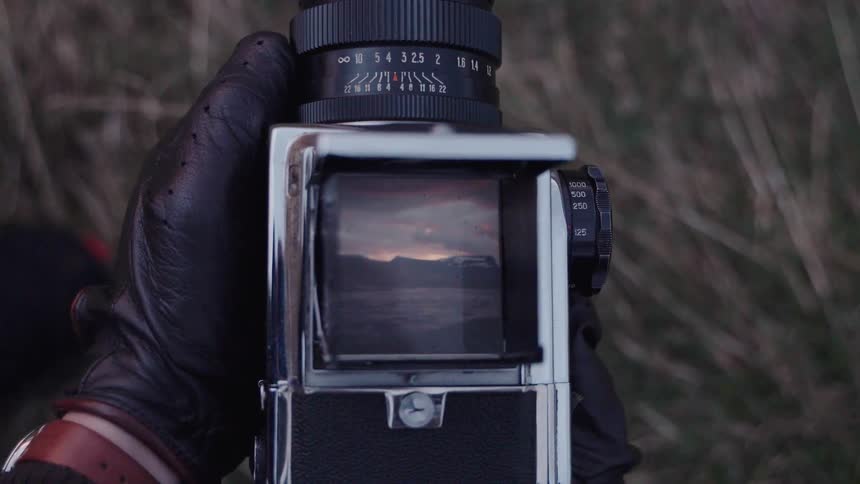 A man wearing a black leather glove holds a vintage medium format camera outdoors viewing a mountain sunset reflected in the ground glass viewfinder