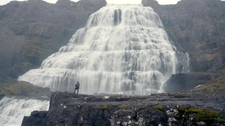 A solitary figure stands on a rocky ledge dwarfed by the immense scale of a powerful multitiered waterfall cascading down a dark mosscovered cliff face under a gray overcast sky