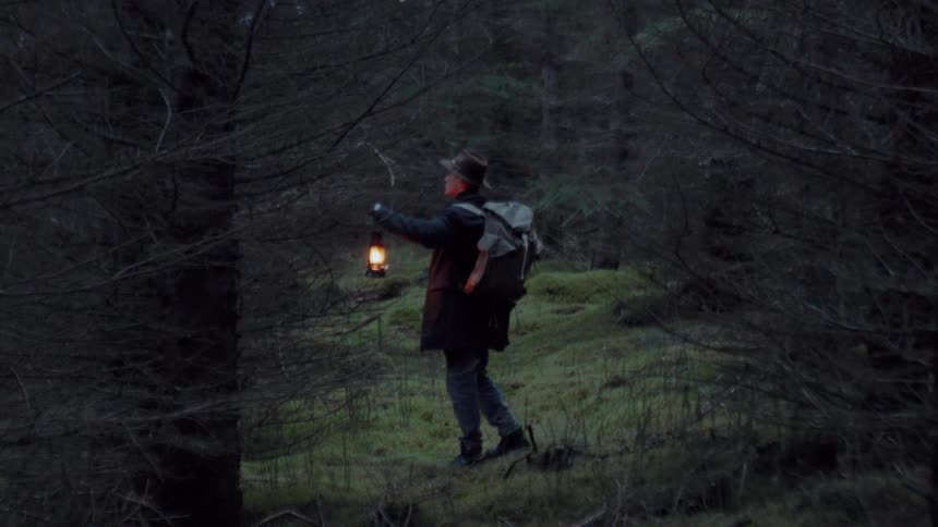 Young adult man wearing a hat and carrying a lamp on his hand while he walks through a dense forest oudoors