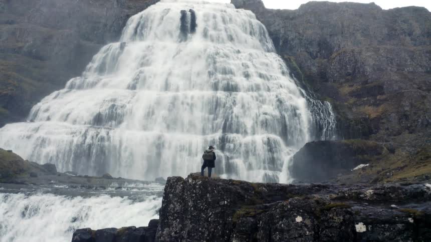 Person standing on a rock facing a large waterfall surrounded by mountains