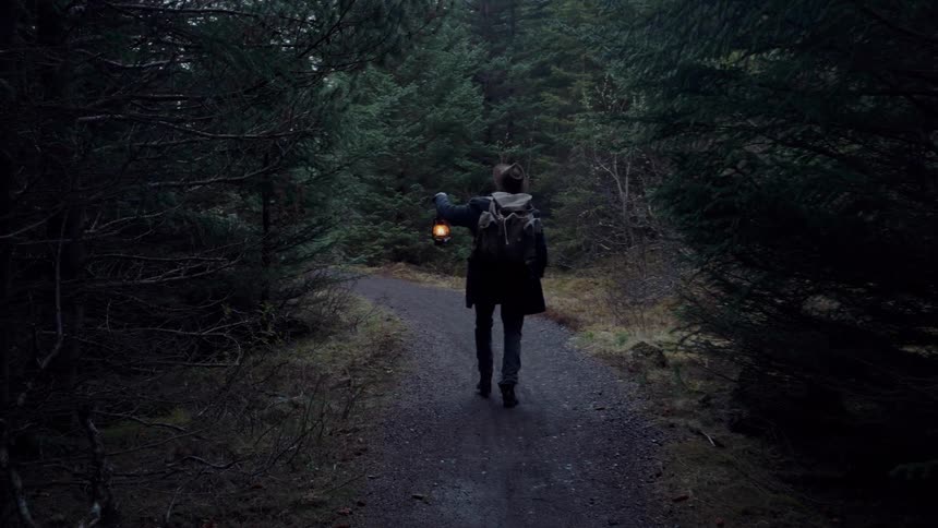 A person wearing a coat a hat and a backpack walks away from the camera down a narrow dark gravel path surrounded by dense forest holding a lit oil lantern in the left hand