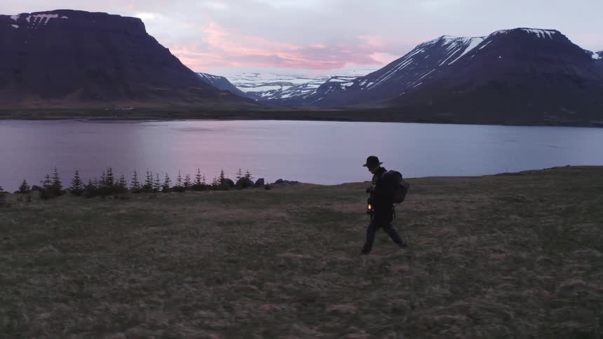 A man hikes past a lake surrounded by mountains while he carries a lantern