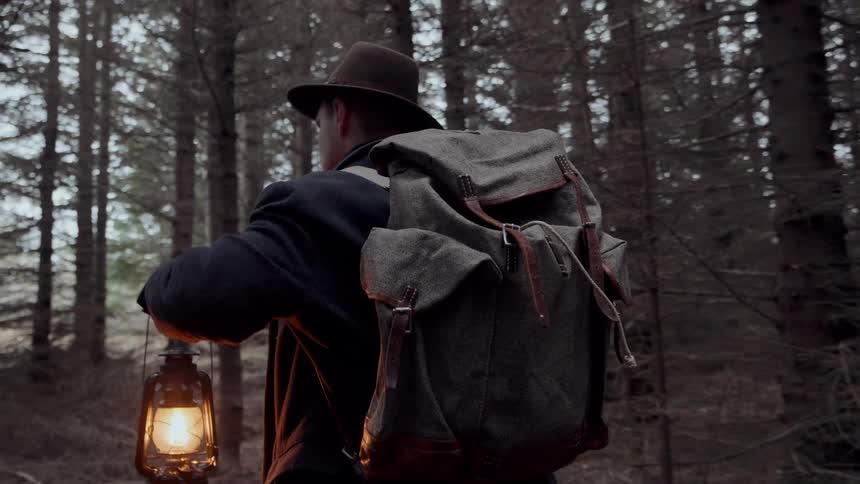 A man wearing a long dark coat and a hat walks through a dense dark evergreen forest carrying a large canvas backpack and a glowing kerosene lantern