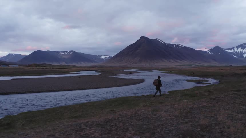 Male hiker carrying a backpack walking next to a river with a mountain range on the background outdoors during dusk or dawn