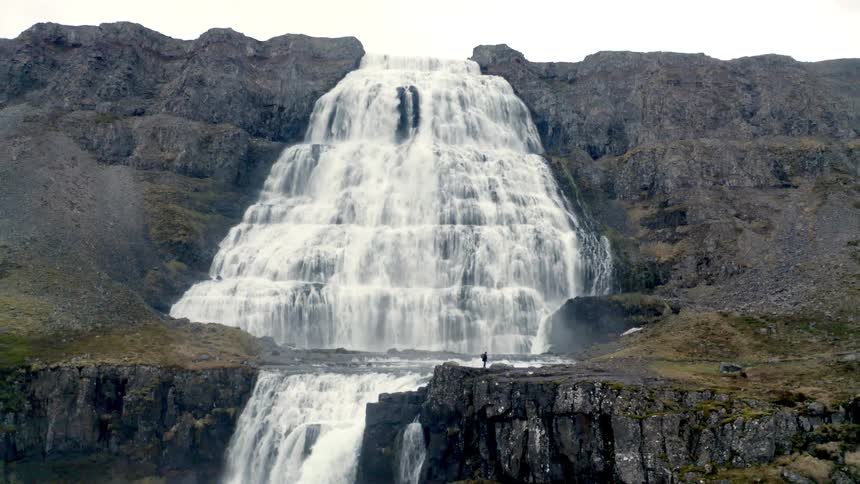 Ahiker is walking with a bright blue sky behind her and a massive tiered white waterfall cascades down steep dark rocky cliffs in a vast remote landscape