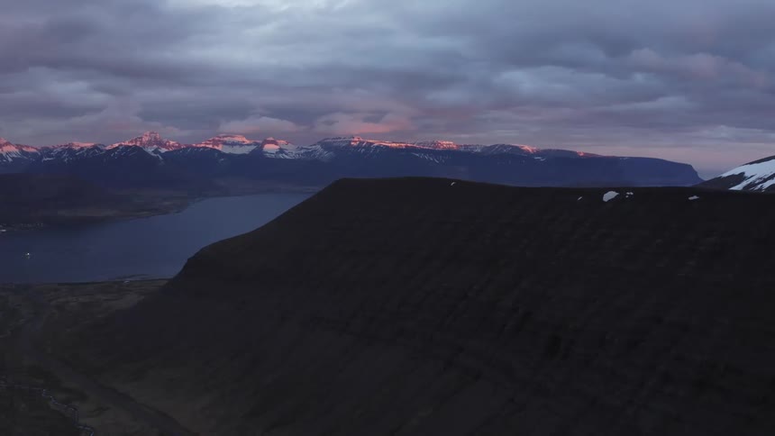 Peak of a mountain next to a lake with a mountain range on the background outdoors during dusk or dawn