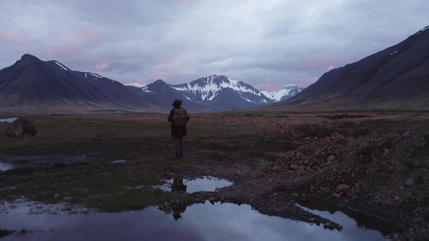 Male hiker wearing a hat and carrying a backpack walking on flat land next to a body of water with a mountain range in the background outdoors during dusk or dawn
