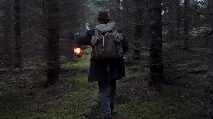 Man wearing a hat and carrying a backpack and a lamp on his hand walking between trees on a forest outdoors