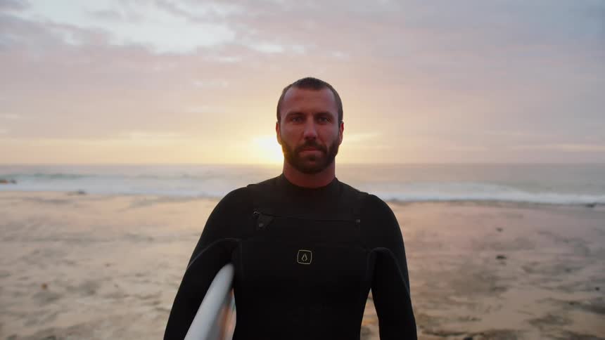 A man in a wetsuit stands on a beach at sunset holding a surfboard
