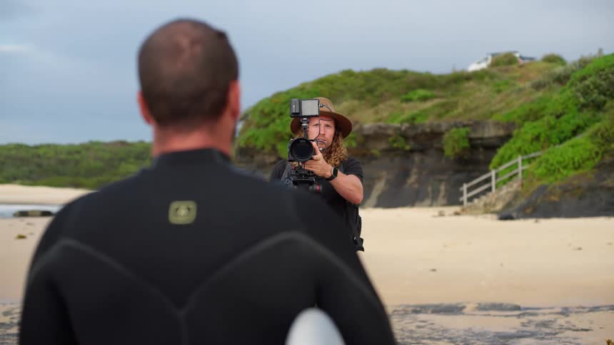 A man in a black wetsuit holding a surfboard is filmed by a videographer with a hat and a professional camera on a tripod at a beach