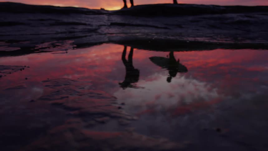 A shallow lowangle shot captures the dramatic red and orange sunset reflected in a waterfilled puddle on a dark rocky surface as the silhouettes of two people one carrying a surfboard walk past above