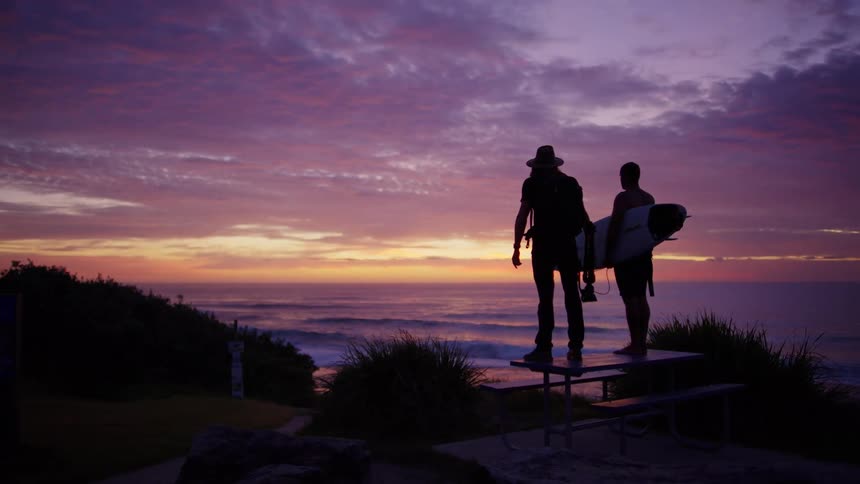 Two people gaze toward the ocean on a beach at sunset with a surfboard in hand