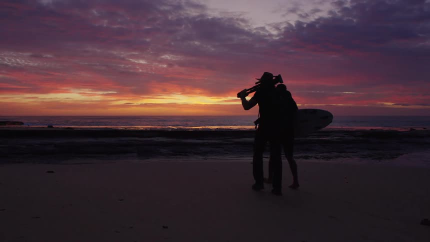 Two silhouetted people walk along a beach carrying a surfboard at sunset
