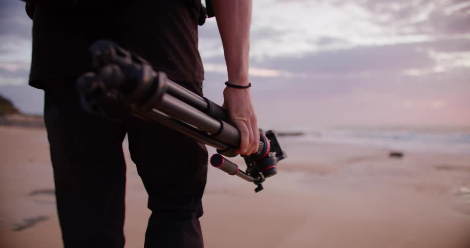 Young adult man wearing black clothes and carrying a backpack and a tripod on his hand walking towards a beach outdoors during dusk or dawn