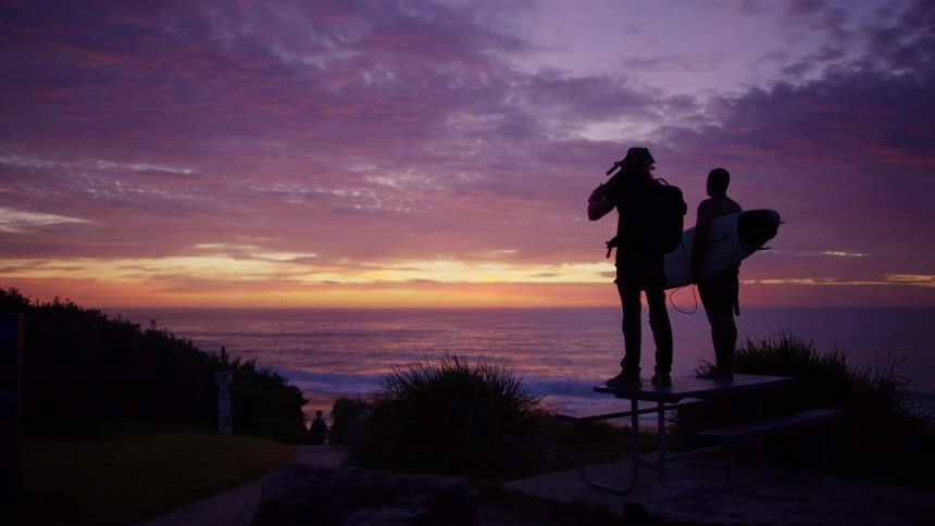 Two men silhouetted on a picnic table watching the seaside sunset one holding a surfboard