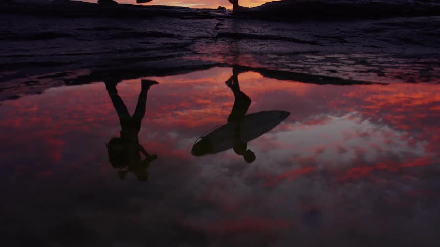 Reflection of a person with a surfboard walking in a tide pool at sunset